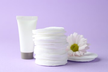 Stack of clean cotton pads, cream and flower on lilac background, closeup
