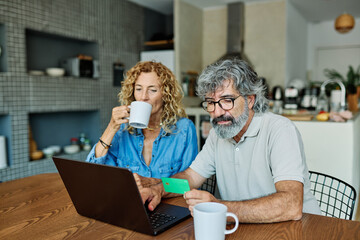 portrait of happy smiling senior elderly couple using a laptop computer and a credit card for online shopping at home, technology and internet use in everyday life concept
