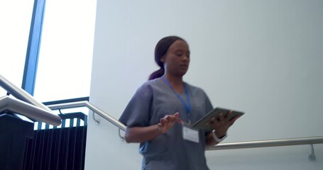 African woman in scrubs checking phone tapping tablet on stair landing showing data to colleague
