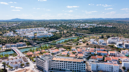 Obraz premium This aerial view showcases Tavira, a town in the Algarve region, with buildings, a river, and the surrounding landscape visible under clear skies