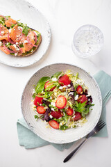 Overhead shot of a strawberry salad on a white plate with a glass of water, a fork, light blue napkin, and another plate with fresh raw salmon on a white background.