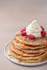 A large stack of pancakes with raspberries, whipped cream, and nuts on a light brown background.