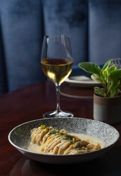 A creamy pasta dish in a fancy bowl with white wine. Dark wood table with blue seats in the background.