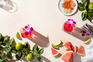Overhead lifestyle shot of fresh limes and leaves, red cocktails with flowers, and slices of watermelon on a light beige surface.