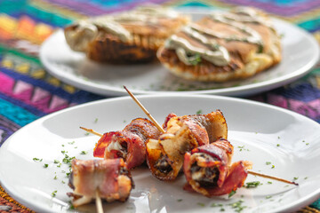 Close-up photograph of casual food made with bacon and meat placed on a white plate with wooden chopsticks with empanadas in the background