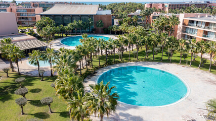 This aerial scene shows the Salgados area of Albufeira, featuring circular swimming pools, palm trees, and nearby buildings amidst a sunny day
