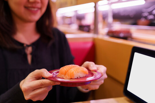 Woman holds a plate of salmon nigiri in a Japanese conveyor belt sushi restaurant.