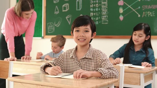 Asian smart boy smiling to camera while student writing answer in answer sheet. Multicultural student doing classwork or test while caucasian teacher checking student homework at classroom. Pedagogy.