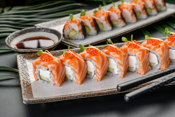 Two plates display sushi rolls made with salmon, rice, and cucumber. Soy sauce sits in a small bowl nearby. Fresh herbs add color to the dish