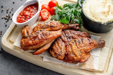 Grilled chicken is placed on a wooden board alongside sauce, fresh tomatoes, greens, and a creamy side dish at a table ready for eating