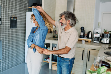 Portrait of happy senior mid aged mature couple prepering meal and having fun dancing and embracing in kitchen at hpme