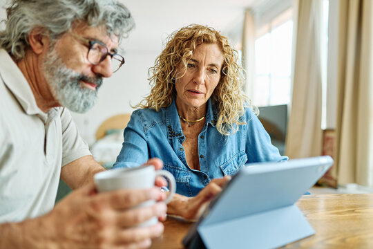 Portrait of a lovely tablet senior mature couple using a laptop together and having fun drinking coffee or tea sitting at a table at home