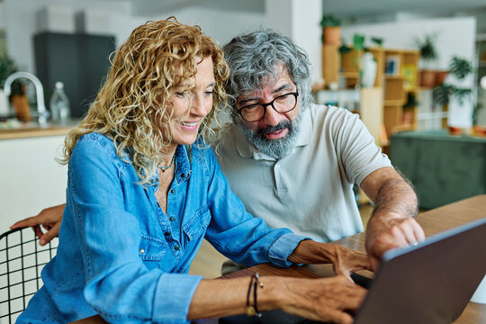 Portrait of a lovely senior mature couple using a laptop together and having fun drinking coffee or tea sitting at a table at home - Powered by Adobe