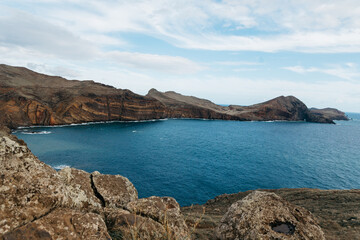 Scenic coastal view featuring rugged cliffs and tranquil blue waters, with rocky foreground and distant hills creating a serene natural landscape