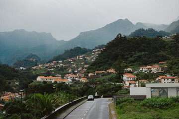 Scenic view of a winding road leading through lush green hills, dotted with charming portuguese houses and surrounded by majestic mountains under a cloudy sky, showcasing natural beauty