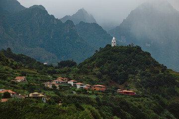 Scenic view of a mountainous landscape featuring lush greenery, quaint houses nestled on hillsides, and a striking white church perched atop a peak, creating a serene atmosphere