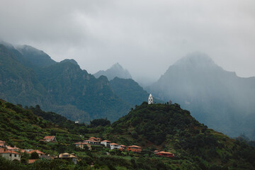 Lush green hills with scattered colorful houses beneath dramatic mountains shrouded in mist, showcasing a serene landscape and natural beauty in a tranquil environment