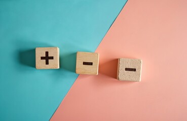 Wooden blocks show plus and minus symbols on a dual toned blue and pink backdrop. These elements symbolize choice and comparison for decision making.