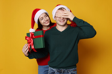 Smiling woman surprising her boyfriend with Christmas gift on yellow background. Happy couple in...