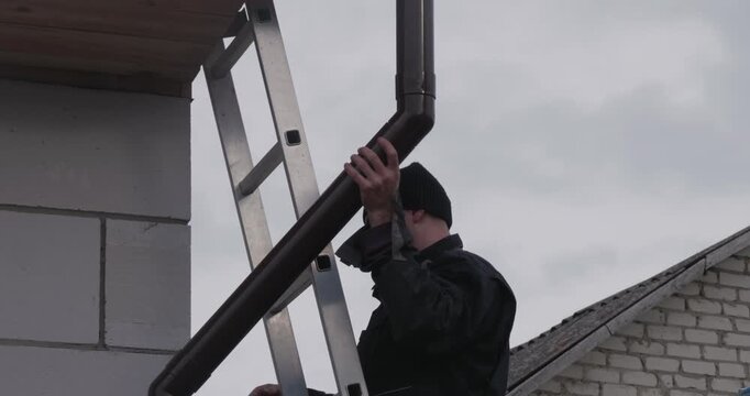A roofer installs a gutter system. The focused craftsman attaches a downspout to the wall of a building under an overcast sky. His confident hands align the metal pipes and secure them to the wall.