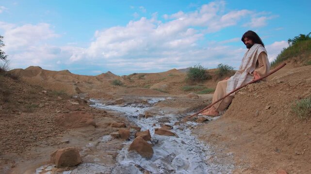 A contemplative scene depicting Christ alone in a desolate and arid landscape. Dressed in traditional white robes, the traveler appears to be resting or praying with a wooden staff at his side.