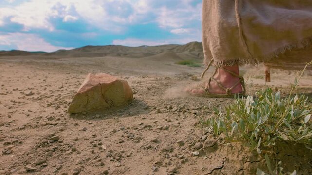 Footsteps of Christ in the wilderness in a sunny day. Slow motion footage of a detailed close-up showing the feet of a traveler clad in ancient rope sandals, standing beside a prominent rock.