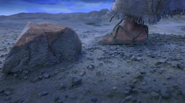 A detailed close-up showing the feet of a traveler clad in ancient rope sandals, standing beside a prominent rock on parched, cracked earth at night.