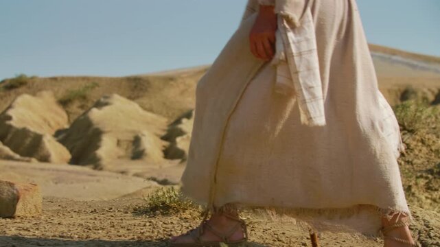 Slow motion shot revealing the feet of a traveler clad in ancient rope sandals, standing beside a prominent rock on parched, cracked earth.