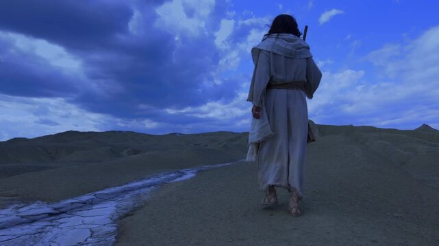 The solitary figure of the Messiah, wearing ancient robe, walks away into the night, crossing a barren and rugged landscape. A dry white stream runs through the foreground, against a dramatic blue sky