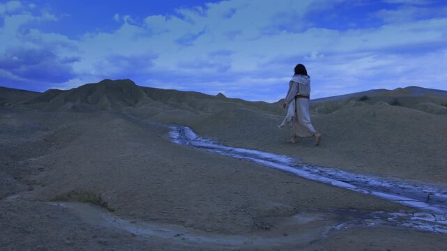 The solitary figure of the Christ walks away into the night, crossing a barren and rugged landscape. A dry white stream runs through the foreground, against a dramatic blue sky