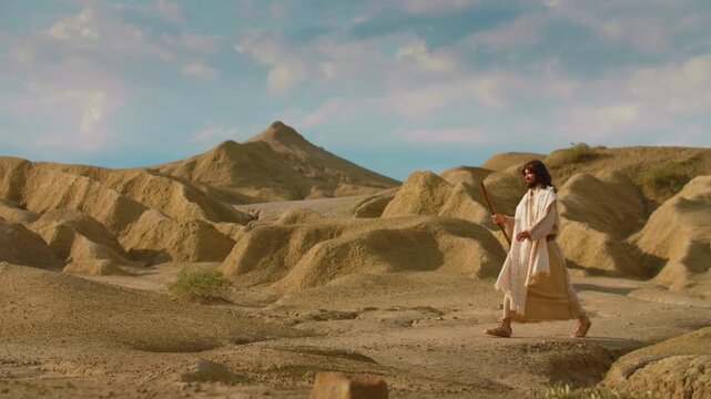 Solitary figure of Jesus dressed in an ancient-style robes walks through a sunlit desert, using a wooden staff for support as they move across the dry, textured ground. Christian and spiritual concept