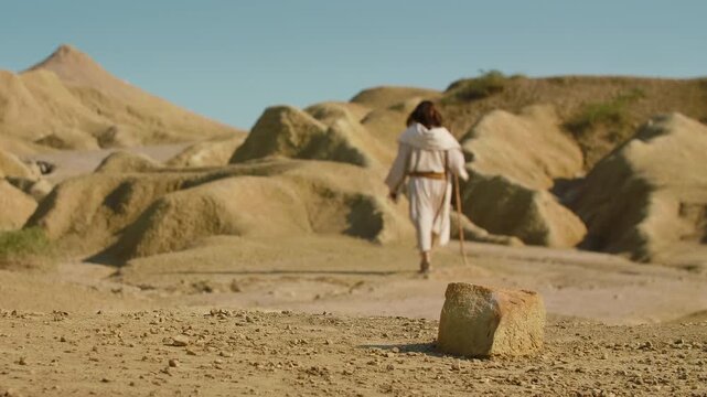 Long Shot of a Solitary Traveler in simple traditional attire crosses an arid, sunlit terrain, using a wooden staff while walking through a quiet and rugged wilderness. Christian and spiritual concept