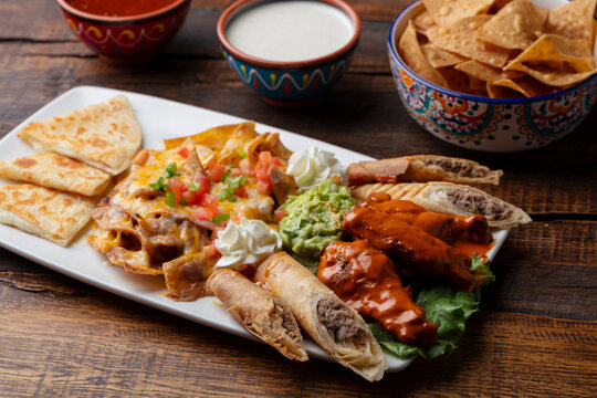 Mexican food appetizer plate with chips, salsa, nachos, taquitos, quesadillas, guacamole, chicken wings with sauce, and sour cream on a white plate. 