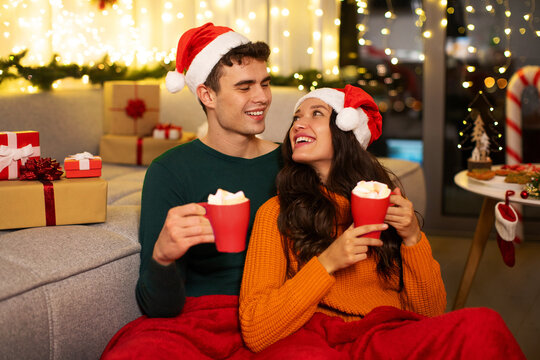 Loving couple holding mugs with cocoa and marshmallows, celebrating Christmas Eve sitting covered with blanket at home. Cozy celebration of winter holidays concept - Powered by Adobe