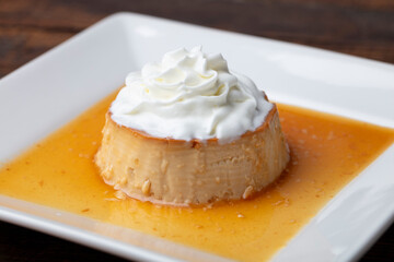 Flan with whipped cream on a small square, white plate and dark background.