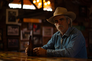 Man With Mustache Wearing Cowboy Hat and Denim Shirt Sitting and Drinking Beer at Bar in Rural Rustic Western Saloon Bar