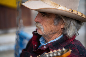 Fototapeta premium Man With Mustache Wearing Cowboy Hat and Red Flannel Shirt Holding Mandolin String Instrument Sitting On Rustic Swing In Front of Old Wooden Wagon Wheel Outside of Rural Saloon Bar