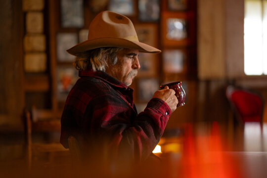 Man Wearing Cowboy Hat Sitting and Drinking Steaming Coffee Out of Red Mug at Table in Saloon Bar With Fireplace in the Background