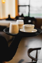 A server holds a black tray with three freshly made, creamy lattes in glasses and a ceramic cup with a saucer in a warm, inviting café environment