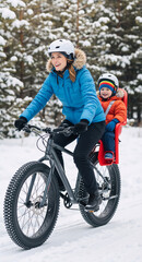 Fototapeta premium Happy mother riding a fat bike with her son in a child seat through a snowy winter forest. Vertical photo of family enjoying outdoor winter activities. Active lifestyle concept