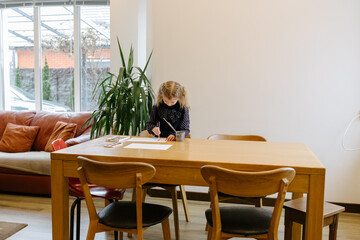 Child drawing at wooden table in modern living room with indoor plant and large windows