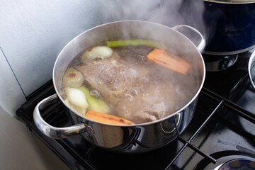 Large stainless steel pot simmering beef, onions, celery and carrot pieces on a gas stove, steam rising as rich homemade broth slowly cooks in a bright family kitchen, bubbles breaking on the surface.
