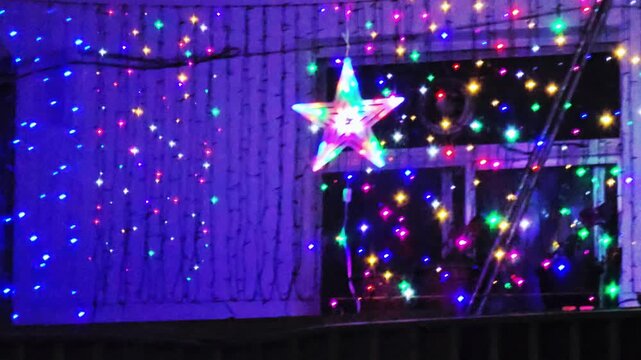 A vibrant outdoor holiday display featuring blue, purple, and multicolor LED string lights cascading down a building, centered around a glowing star decoration.