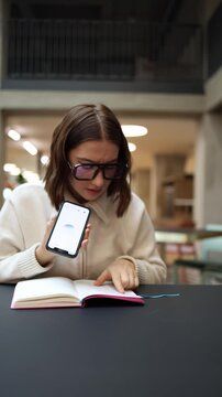 Vertical screen young woman holds her phone with Чат GPT open and studies her notes while pointing at the page trying to clarify details through active voice interaction in a bright workspace