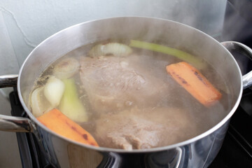 Large stainless steel pot simmering beef, onions, celery and carrot pieces on a gas stove, steam rising as rich homemade broth slowly cooks in a bright family kitchen, bubbles breaking on the surface.