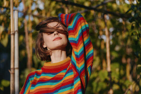 Woman portrait in a rainbow sweater outdoors, relaxed and basking in warm sunlight, short hair framing face and green nature background for cozy lifestyle, fashion and seasonal mood.