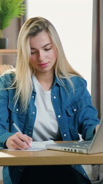 Adorable satisfied skilled 25-aged girl in casual clothes sitting at her workplace in working home room and reading report on computer nd writing down important notes on sheet of paper