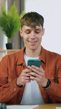 Good-looking positive carefree 23-aged male person in orange shirt sitting at his workplace with laptop in home office and chatting with friends on smartphone