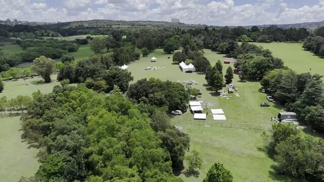 Drone orbits to the right over fairgrounds with large white tents next to large field boxed in by a forest on partially cloudy day in Randburg, Johannesburg, South Africa