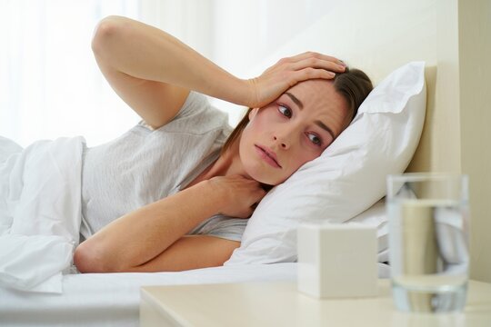 Unwell woman in bed holding head and neck, suffering from headache and discomfort